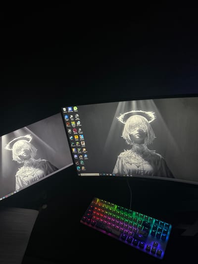 The image shows a dual-monitor setup with a black desk and a mechanical keyboard. The monitors display a black and white wallpaper of an angelic figure with a halo and obscured eyes. The left monitor shows a portion of the figure and some desktop icons. The right monitor displays the full figure and more desktop icons, including various game launchers and applications. A book is visible on the left side of the desk, and the mechanical keyboard, which has RGB lighting, is positioned in the lower right corner. The environment is dimly lit, with the primary light source appearing to be the glow from the monitors. The scene is indoors, likely in a home office or gaming setup. There is no indication of the location being in Staromyshastovskaya, Russia.