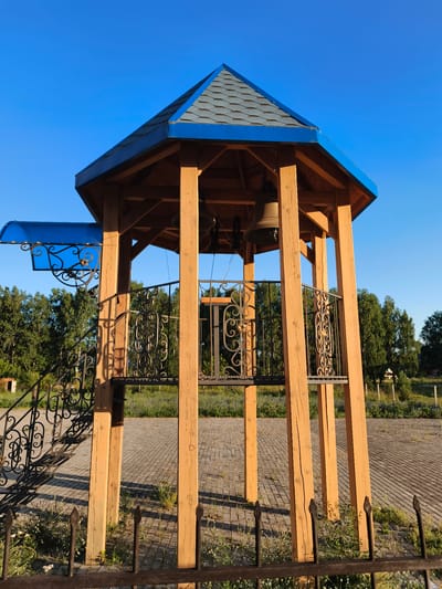 A bell tower stands on a paved area near a wooded area. The bell tower is octagonal in shape with wooden posts and a blue-shingled roof. There are two bells hanging inside the tower, and ornate metal railings surround the upper platform. A blue awning is attached to one side of the tower, above a set of metal stairs. The sky is clear and blue. The area appears to be in the city of Aya, Russia.