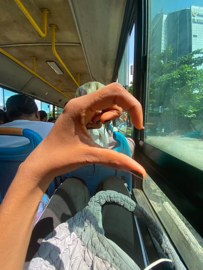 A first-person perspective shot captures a hand reaching out within a bus. The hand, with dark nail polish, is positioned in the foreground, obscuring some of the view. Through the bus window, a reflection shows trees and a building, suggesting an urban environment. Inside the bus, yellow grab rails are visible along the wall. Passengers are seated behind the hand, with one person's head and shoulders visible in the lower part of the frame. A light blue seat is also partially visible. The overall lighting indicates it is daytime. The scene is set on a bus, likely in Ikoyi, Nigeria, based on the location context.