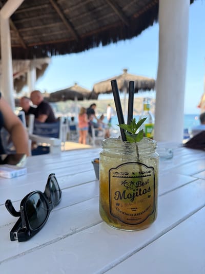 A refreshing mojito in a jar with mint sprigs and black straws sits on a white wooden table. The label on the jar reads "Best Mojitos & Beach Party" and "Cocktails Artesanos Since 1988". Sunglasses are placed on the table next to the drink. In the background, beach umbrellas and loungers are visible, suggesting a sunny beach setting. People can be seen relaxing in the background. The image appears to be taken during the daytime, with clear skies indicating good weather. The location is identified as Xàbia / Jávea, Spain.