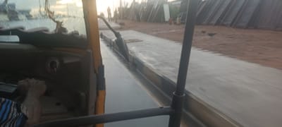 The image, taken from inside a moving yellow vehicle, shows a wet, paved road in Umuahia, Nigeria. A strip of bare ground runs parallel to the road. On the right of the image, a pile of long, dark objects are stacked, possibly construction materials. Two figures stand in the distance. Inside the vehicle, a hand in the lower left grasps a control. A sticker is visible on the dashboard. The road and objects outside the vehicle are blurred, suggesting motion. Two chickens are visible.