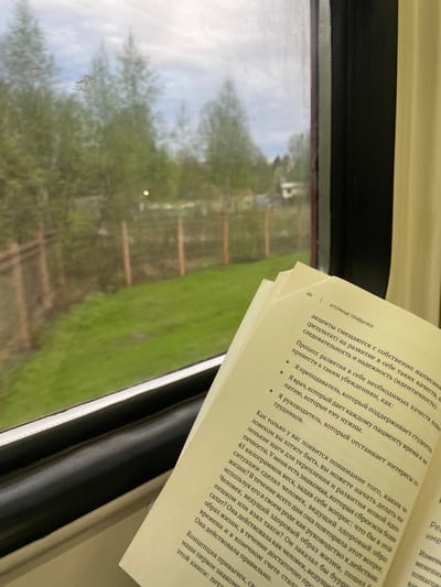The image shows an open book on a surface, likely inside a train or bus in Ashukino, Russia. The book's pages are visible, with text in Russian. The window behind the book shows a blurry view of trees and sky outside. The text on the open book page is about personal development.
