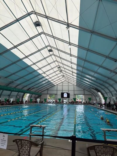 This image depicts an indoor swimming pool facility in Chamblee, United States of America. The facility is housed under a large, vaulted or A-frame structure featuring a roof composed of alternating translucent white and opaque light blue panels supported by a metal framework. Multiple dome-shaped lighting fixtures are suspended from the ceiling, alongside several rectangular ventilation units.
The central feature is a multi-lane swimming pool filled with clear blue water. Approximately 8-10 lanes are delineated by green and yellow lane lines. Several individuals are present in the pool; some are actively swimming laps, while others are standing or congregating near the ends.
Along the far side of the pool, bleachers filled with spectators are visible. Above the spectator area, green triangular pennants are suspended. A large digital screen, mounted on the far wall, displays text that reads "BIG NIGHT OUT" along with graphics, suggesting an event is taking place. To the right of the screen, an arched opening in the structure provides a view of an outdoor area with trees and a yellow construction vehicle.
Along the lower interior curve of the ceiling structure, a continuous green, fabric-covered ventilation duct is visible. In the immediate foreground, a white starting block is positioned at the edge of the pool. Partially visible are two empty, brown chairs with a geometric lattice pattern on their backs. A white sign with black text is posted in the foreground, stating: "NO Photography of any kind behind the blocks." Electrical cables or cords are visible on the concrete deck near the chairs and starting block.