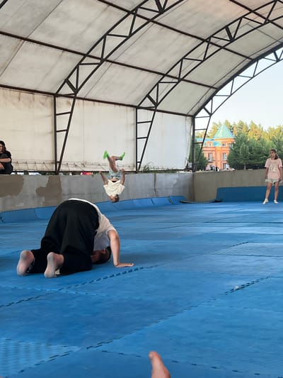 The image captures an outdoor training session, likely a martial arts or parkour practice, taking place under a simple open-sided structure with a metal frame and translucent roof. The ground is covered with blue interlocking mats, commonly used for such activities.
Several individuals are present. In the mid-ground, a person is mid-air, performing a jump or flip, dressed in a white t-shirt and patterned shorts with bright green socks. Another person is on the blue mats in the foreground, in a crouched or rolling position, wearing a white t-shirt and black pants. Further in the background, another individual stands, wearing a pink t-shirt and shorts. A person's hand and arm are visible in the far right foreground, suggesting another participant or observer.
The scene appears to be set during daylight, with the sun casting shadows, indicating it's likely daytime, possibly late afternoon given the angle of the light. The background shows trees and a building with a distinctive green roof and ornate brickwork, which is identified as being in Krasnoturansk, Russia. The overall atmosphere seems active and focused, with the participants engaged in physical training. There is no visible text in the image.