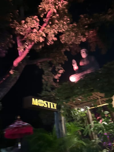 The image shows an outdoor night scene in Pererenan, Indonesia, near the restaurant "Mostly." The view is obscured by trees and foliage. Illuminated hanging lamps are visible above. A sign for the restaurant is present and lit. Additionally, a red, round object is partially visible near the sign. The scene appears dark and lush with vegetation.