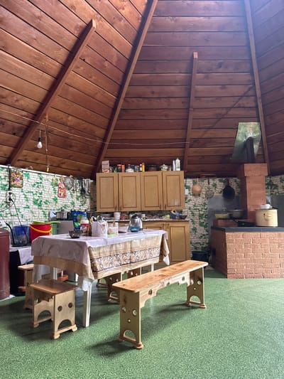 The image depicts the interior of a rustic kitchen with a high, sloped wooden ceiling. The walls are partially covered with green and white brick-patterned wallpaper. In the center, a table is set with a patterned tablecloth, surrounded by wooden stools and benches. To the left, there are kitchen cabinets above a counter with various kitchenware, including pots, a kettle, and bowls. A red bucket and a blue bucket are also visible. To the right, a large brick oven with a metal chimney occupies a significant portion of the space. The floor is covered with a green, textured material resembling grass. The scene suggests a functional, lived-in kitchen, possibly in a dacha or rural home in Yabogan, Russia, given the presence of a traditional brick oven and the overall rustic aesthetic. No people are present in the image.