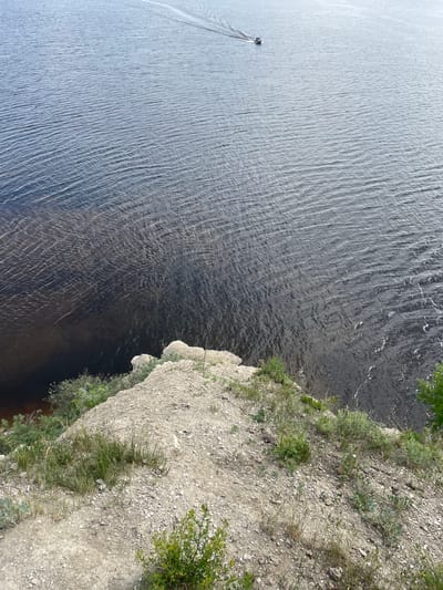 From a high vantage point, the image presents a vast expanse of water alongside a rugged, rocky cliff. The scene is illuminated by bright daylight, indicating clear weather conditions.

The left side of the frame is dominated by a large body of water, possibly a river or lake, featuring numerous ripples across its surface that suggest gentle movement or wind. The water's color varies, appearing lighter towards the upper left, potentially reflecting the sky, and gradually becoming darker towards the bottom right. In the upper left quadrant of the water, a small, dark-colored boat is seen traversing the surface, leaving a distinct white wake trailing behind it.

The right side of the image is occupied by a steep, earthy cliff or hillside. The ground is composed of light-colored rocks and soil, with sparse patches of green vegetation, including grasses and small bushes, growing along its edge and clinging to the slope. The perspective looks sharply downward, emphasizing the height of the viewpoint above the water.

This natural landscape, featuring a moving boat and a vegetated cliff next to a body of water, is situated near Пятый километр, Russia. There is no visible text in the image.