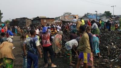 The image displays an outdoor scene in Gisenyi, Rwanda, featuring a large group of approximately 50 to 70 individuals engaged in the clearing of a substantial waste accumulation. The central foreground and midground are dominated by a dark, irregular mound of debris composed of earth, organic matter, and visible discarded items such as plastic bottles and other refuse.

Numerous individuals, primarily adults and youth, are actively working on or around this waste pile. Actions observed include sweeping, gathering, and moving material using rudimentary tools, specifically bundles of sticks fashioned into brooms. Several participants are wearing high-visibility yellow vests, indicating an organized effort. Individuals are dressed in diverse casual clothing.

The environment consists of an unpaved, dirt ground. In the background, a dense cluster of single-story residential structures constructed from wood planks and corrugated metal sheets extends across the frame, signifying a densely populated urban or semi-urban area. The sky is light grey and appears overcast. An electricity pole with overhead lines is visible on the right side of the frame in the background.
