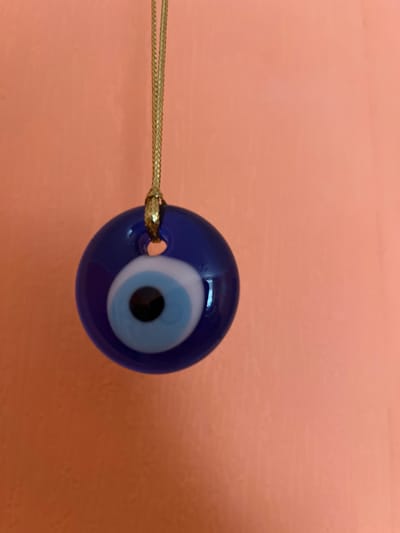 An evil eye amulet, characterized by concentric circles of blue, white, and black, hangs from a thin gold chain against a textured, peachy-pink wall. The amulet is round and appears to be made of glass. The gold chain is braided and has a knot near the top where it attaches to the amulet. The wall has a slight texture, suggesting it might be painted plaster or a similar material. The lighting appears to be natural, casting subtle shadows on the wall behind the amulet. The scene is static and depicts a single object in a simple indoor setting. There is no indication of a city or geographical location.