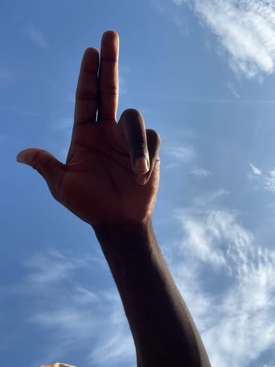 A dark-skinned human hand and forearm are positioned against a clear blue sky featuring scattered white cirrus clouds, observed in Gbaza, Nigeria. The index and middle fingers are extended upwards and held together, while the ring finger, pinky finger, and thumb are bent. The hand and lower arm are largely silhouetted against the bright sky, with some light visible along the edges of the extended fingers. Minute specks are present in the sky background.
