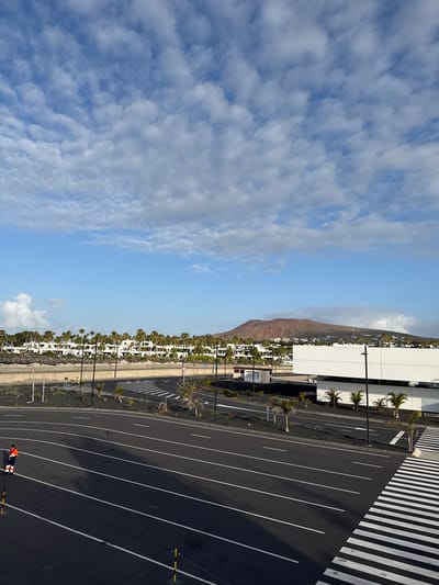 The image shows a view over Yaiza, Spain, a municipality in the Canary Islands. A brown-colored mountain rises in the background. The sky is blue with some scattered clouds. A dense row of white buildings stretches across the midground, interspersed with trees. The foreground consists of an asphalt parking lot with white painted lines, a crosswalk, and utility poles. An individual wearing a safety jacket can be seen in the lower-right corner of the image.