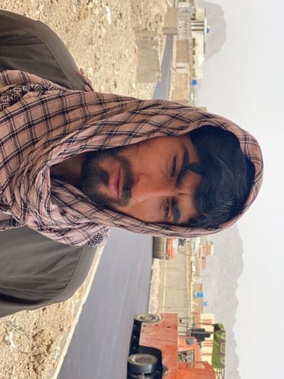 A man wearing a tan and brown plaid headscarf stands beside a newly paved asphalt road in Kandahar, Afghanistan. Buildings line the road in the distance, and mountains are faintly visible in the background. Construction equipment sits at the edge of the road, indicating ongoing construction activities.