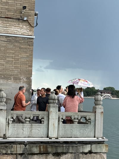 The image depicts a crowded outdoor scene on a stone balustrade overlooking a large lake in the Haidian District of Beijing, China, specifically within the Summer Palace (Yiheyuan) complex, likely facing Kunming Lake towards Nanhu Island.
In the foreground to the left, a weathered light-brown brick wall is visible. Mounted on this wall are two security cameras: a white dome camera facing downwards and a silver bullet-style camera extending horizontally, both connected by visible conduits and wiring, some of which show signs of rust. Below the brick, the wall transitions to a lighter, stained stone or concrete base.
A grey stone balustrade, intricately carved with ornate pillars featuring vase-like tops and decorative panels with cloud or scroll motifs, spans across the lower half of the frame. Behind this balustrade, a group of approximately eight people, primarily adults, are gathered.
From left to right along the balustrade:
* A Caucasian male in an orange short-sleeved shirt with a black shoulder bag is speaking to another man. His left hand is slightly raised in a gesture.
* Next to him, an Asian male in a dark grey short-sleeved shirt stands with his back partially turned, looking towards the lake.
* Partially obscured behind them is an individual in a white hooded garment.
* Further to the right, an Asian female in a light pink short-sleeved top holds a colorful umbrella with a floral pattern over her head, using both hands to photograph or video the distant scenery with a smartphone.
* Other individuals are visible to her left and right, also observing the view, with at least one more person holding a smartphone.
The background features a vast expanse of calm, greenish-blue water, which is Kunming Lake. On the far side of the lake, a traditional Chinese architectural complex stands on a low, tree-covered landmass, consistent with Nanhu Island. The prominent building has dark roofs, white walls, and a distinctive arched entrance or bridge leading up to it, surrounded by lush green trees. The sky above is overcast with dark grey clouds, indicating cloudy weather.