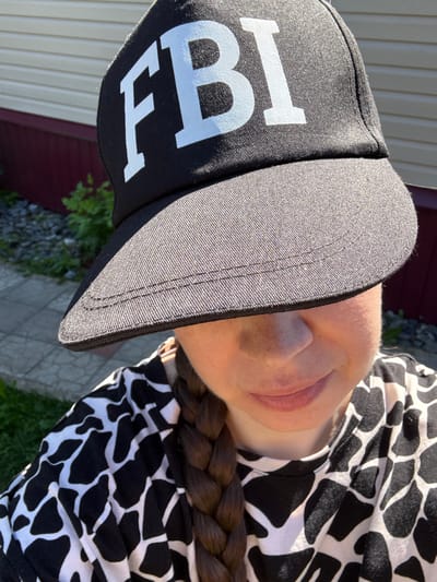 A young Caucasian woman is captured from the chest up, wearing a black baseball cap with white "FBI" lettering. The cap's brim casts a shadow over her eyes. She has a single braid of brown hair extending over her shoulder and is wearing a black and white patterned shirt. The background shows the exterior of a building with horizontal siding and a dark red lower portion, adjacent to a gravelly area with green foliage and a paved walkway. The scene is lit by direct sunlight.