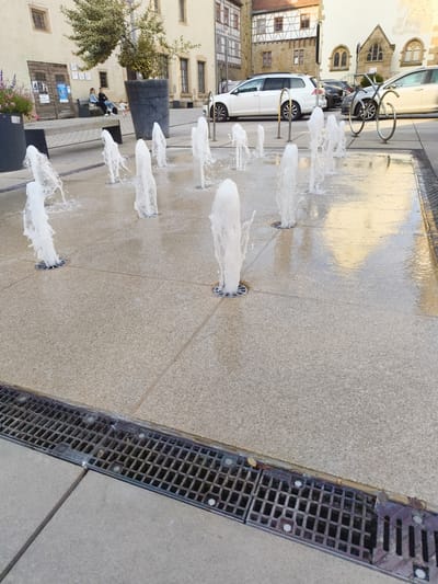 The image captures a public square in Neuenstadt am Kocher, Germany, featuring an active water fountain installation. In the foreground, several water jets erupt from circular grates set into a textured, light-colored pavement. The water sprays upwards, creating a frothy, dynamic effect. A metal grate drain runs along the bottom left edge of the frame. In the middle ground, a bench with two individuals seated and a small child is visible, positioned near large planters and a building with traditional German architecture. Behind them, several cars are parked. The background reveals more buildings, including one with a distinctive arched window. The overall scene suggests a pleasant, sunny day in a European town center.