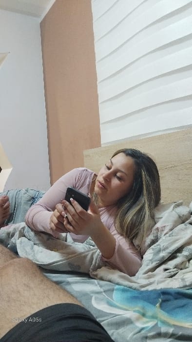 The image depicts an indoor scene, stated to be in a bedroom in Oreshene, Bulgaria. A female individual with light brown and blonde hair, wearing a light pink long-sleeved top and light blue denim jeans, is lying on her left side on a bed. Her head is resting on a pillow against a light-colored wooden headboard. She is holding a black smartphone with both hands, focusing her gaze downward at its screen. Her fingernails feature white tips. To the left, in the foreground, the upper right thigh and knee of a second individual, a male with visible leg hair and dark shorts or underwear, are partially visible. The bed is covered with a patterned duvet, grey with a light blue floral design, and a lighter-colored blanket is draped over parts of the bedding and the female subject's lower body. The background features a light brown wall on the left and a white wall with horizontal, corrugated-like paneling on the right. The image contains a "Galaxy A05s" watermark in the bottom left.