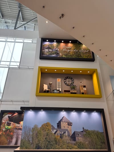The image shows three displays attached to a wall in what appears to be a public building, likely in Remscheid, Germany. The leftmost display is a black-framed picture showcasing a landscape, with the text "Unser Bergisches Land" visible on the print. The middle display is a yellow-framed box filled with various objects, including a clock, small shelves containing items, a vintage desk lamp, and an open book. The rightmost display is a black-framed picture that seems to have two separate scenes stacked vertically: the upper section depicts a castle or large stone building surrounded by trees under a cloudy sky, while the lower section reveals a cable car or suspension railway over a body of water. Below the displays, there are window blinds. The surrounding environment includes architectural elements such as supporting pillars and a ceiling with metal beams and skylights, suggesting the building is a modern structure.