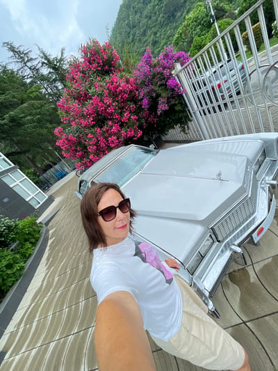 A woman in sunglasses and a white t-shirt takes a selfie in front of a vintage silver car. The car's hood and grill are visible. Behind the car, there's a large bush with pink and purple flowers and a metal fence. In the background, there are trees and a cloudy sky. To the left, part of a modern building with large windows is visible, and green bushes line the walkway. The ground is paved with interlocking stones that appear wet. The scene is likely outdoors in a temperate climate. The presence of a vintage car and the architecture suggest a location with a mix of historical and modern elements.