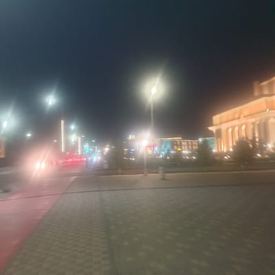 At night in what appears to be the city of Turkistan, Kazakhstan, a street scene is illuminated by streetlights. A wide sidewalk composed of interlocking paving stones extends into the foreground, with a red painted lane closer to the street. Buildings with illuminated windows can be seen across the street. The sky is dark and clear. Cars with headlights are visible. No people are clearly visible, though there may be blurred figures on the sidewalk.
