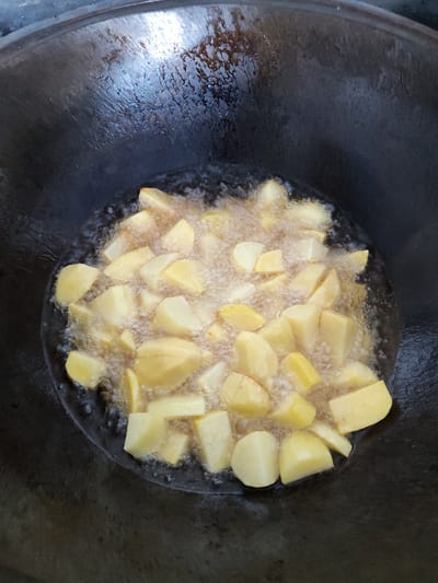 This is a top-down, close-up shot of chopped potatoes being deep-fried in a large, dark, wok-like pan. The pan is partially filled with bubbling hot oil, and the potatoes are submerged, cooking and releasing steam. The potatoes are cut into irregular, bite-sized pieces, with some showing a golden-brown hue starting to form, indicating they are browning. The oil is frothy and active, with many small bubbles surrounding the potato pieces. The pan's surface is dark and appears to be seasoned cast iron, with some textured markings visible on the un-oiled parts. The lighting suggests it could be daytime, possibly indoors given the focus on the cooking process. The scene is entirely focused on the food preparation, with no other objects or people visible. The location context of Yalangach, Uzbekistan, hints at a traditional culinary setting where deep-frying is common. There is no visible text in the image.
