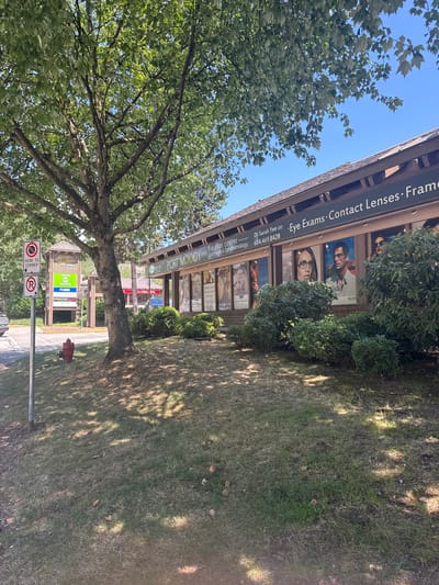 A treelined outdoor scene in Port Moody, Canada, depicts a commercial building with a prominent sign reading "PORT MOODY EYE CARE CENTRE". The sign also lists services such as "Optometry & Ophthalmology" and contact information "Dr. Sarah Yee OD 604.469.8428". Below the sign, large posters showcase models wearing eyeglasses, including one with the "BOSS" logo. Lush green bushes flank the storefront, and a large deciduous tree with a thick trunk and sprawling branches shades the foreground. In the background, a smaller building with a peaked roof and the "Heritage Mountain" logo is visible, along with a "Save On Foods" sign. Two red and white regulatory signs are mounted on a pole to the left of the frame, one indicating "NO PARKING HERE TO CORNER" and the other a "NO RIGHT TURN" symbol. A red fire hydrant is situated on a grassy slope near the base of the tree. The ground is covered in dry grass with scattered fallen leaves, and dappled sunlight filters through the tree's canopy. The sky is clear and blue.