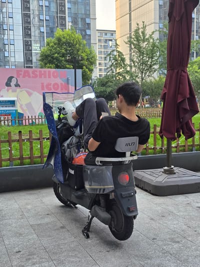 A young man sits on an electric scooter, facing away from the camera. He is wearing a black t-shirt and black pants, with white sneakers. The scooter is parked on a paved sidewalk next to a grassy area with a wooden fence. In the background, there is a large building with many windows and some trees. A red umbrella is partially visible in the upper left corner.

The scooter has a gray body and a blue fender. There is a red taillight on the back of the scooter. A plastic shield covers the front of the scooter. A sign with the words "FASHION ICON" in large pink letters is visible next to the scooter. The sign also features an illustration of a person's face. Some Chinese text is also visible on the sign, including "分类杭州更美" (Classification makes Hangzhou more beautiful). The image appears to have been taken during the daytime. The overall scene suggests an urban setting, possibly a street or a public area.