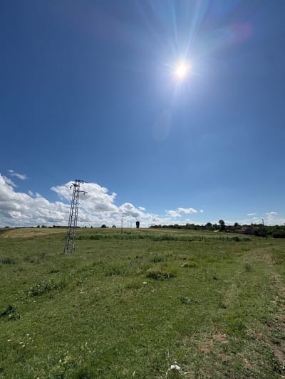 The image presents a vibrant, sunlit landscape, likely captured in the rural environs of Tarnak, Bulgaria. The scene is dominated by a brilliant blue sky, mostly clear but with scattered white, puffy cumulus clouds adding texture to the horizon and mid-sky. A dazzling sun, emitting strong starburst rays and a lens flare effect, is prominent in the upper left quadrant, indicating bright daylight, probably midday or early afternoon.

Below the expansive sky, a vast green field stretches across the mid-ground and foreground. The grass appears natural and somewhat wild, suggesting a pasture or undeveloped land, with varying shades of green and some brown patches.

A significant man-made structure, a metal electricity pylon or transmission tower, stands prominently in the mid-ground, extending from the lower center towards the right. Multiple power lines are visible, connecting to the pylon and stretching across the sky, suggesting an active electrical grid. Further in the distance, another, taller pole or smaller pylon is visible near the horizon to the right. Along the distant horizon line, faint silhouettes of trees and what appear to be buildings or houses indicate a small settlement or village. No specific activity or individuals are visible within the frame, emphasizing the quiet, open nature of the countryside. There is no visible text in the image.