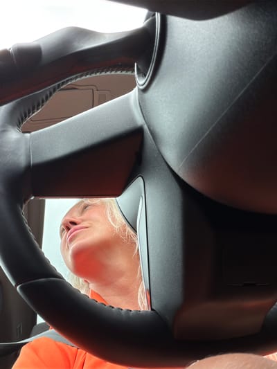 The image depicts a low-angle, close-up shot of a person inside a vehicle, looking upwards. The foreground is dominated by the steering wheel of a car, which is black and appears to be made of leather with visible stitching. The camera angle is from the driver's perspective, looking through the steering wheel.
A woman with blonde hair is visible in the background, her head tilted back as she looks upwards, possibly at the sky or something outside the car. She is wearing an orange garment, possibly a safety vest or jacket, and a portion of her neck and chin are visible. Her facial features, including her nose and lips, are discernible.
The interior of the car is dark, with the exception of a bright, overcast sky visible through the windshield, creating a stark contrast. Shadows are cast by the steering wheel onto the interior surfaces. The specific location in the city of Salford, United Kingdom, is not identifiable from the visual information provided in the image. The scene appears to be captured during daylight hours.