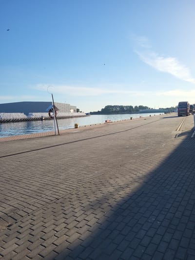This is a photograph of a harbor or dock area in Gdańsk, Poland, taken during the day. The scene is dominated by a paved walkway made of interlocking bricks, which occupies the right side of the frame and extends into the distance. A shadow from an unseen object creates a diagonal line across the walkway.

To the left of the walkway is a body of water, likely a canal or river, with several yellow buoys marking its edge. Along the water's edge, there are rows of white objects, possibly bags of sand or construction materials. A white building with a gray roof is partially visible behind these objects. A red and white life preserver is mounted on a pole extending over the water.

In the background, a line of trees borders the water. Further in the distance, along the far edge of the water, a few buildings and structures can be seen, including a red and silver truck parked on what appears to be a road. The sky is blue with some wispy white clouds. A few small dark shapes, likely birds, are visible in the sky. The lighting suggests it is daytime, possibly late morning or early afternoon, with clear weather. There is no visible text in the image.
