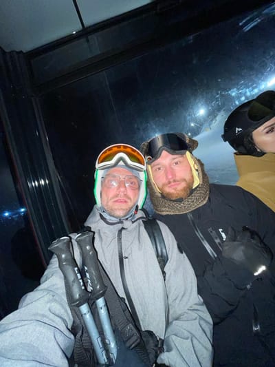An image taken at night depicts three individuals inside a ski lift cabin, likely a gondola, at the Jahorina ski resort in Bosnia and Herzegovina.
In the foreground, an adult male on the left faces the camera, wearing a light grey zippered ski jacket, a white helmet with a green trim, and orange-lensed ski goggles pushed up onto the helmet. He also wears clear-rimmed eyeglasses and has a beard. He holds two grey ski poles with black grips, bearing the "LEKI" brand name, upright in front of him. A black backpack strap is visible on his right shoulder.
To his right, a second adult male looks towards the camera, dressed in a black zippered ski jacket. He wears a textured brown knitted balaclava or neck gaiter, with dark ski goggles pushed up onto his forehead. He also has a beard. A black ski glove is visible on his left hand.
Partially visible on the far right is the profile of a third individual, likely an adult female, wearing a black ski helmet and a light brown jacket. Her right eye and part of her cheek are discernable.
The background, visible through the cabin windows, shows a snow-covered ski slope illuminated by artificial lights, indicating night skiing conditions. The overall scene is dark, typical of nighttime in a mountain environment.