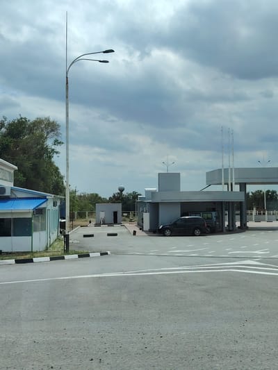 The image shows a checkpoint or entrance area. In the foreground is a paved road with white lane markings and a speed bump. To the left is a white building with a blue roof and windows. A tall pole with two streetlights stands near the building. In the middle ground, there is a grey building with a canopy where two vehicles are parked, a dark SUV and a van. There are also three flagpoles behind the canopy. Further back, there is a small grey building with two doorways and a water tower visible above the trees in the distance. The sky is overcast with clouds. The scene is set in an outdoor, possibly industrial or administrative area, located in the city of Eksperimental'nyi, Russia.