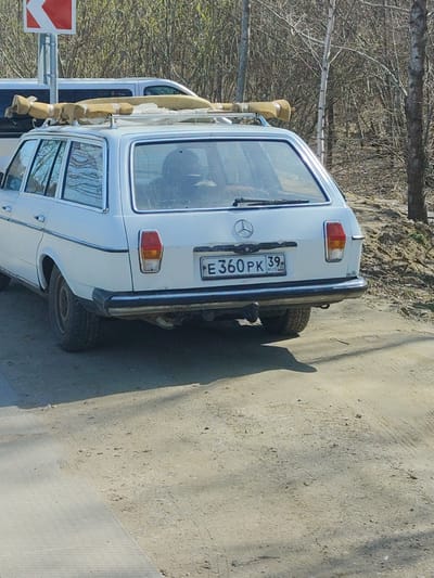 A white Mercedes-Benz station wagon with a roof rack containing wrapped and secured objects is parked on a dirt road next to a paved surface. The car has a Russian license plate that reads "E360PK 39 RUS". The background features a road sign, bare trees, and a grassy embankment. The location is likely in a rural or suburban area in Primorye, Russia, considering the presence of Russian license plates and the surrounding natural environment. There are no visible people directly interacting with the car.