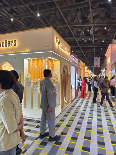 The image depicts an indoor scene within an exhibition hall, located in the city of Jaipur Municipal Corporation, India.
The scene presents a bustling aisle lined with exhibition stalls, characteristic of a trade show. In the immediate foreground, to the left, a person wearing a light-colored, short-sleeved top and blue jeans stands with their back mostly to the viewer, hands clasped. Central to the mid-foreground is a man in a light grey suit and black athletic shoes, standing with his back to the camera, intently observing the display of the adjacent stall.
The prominent stall on the left is labeled "Saraf Jewellers" with illuminated gold lettering and a diamond-shaped logo. The booth is predominantly white with gold trim and features several large glass display windows showcasing various silver and gold necklaces and other jewelry pieces on busts and pedestals. The number "1815-16" is visible on the booth's fascia. The stall's design incorporates scalloped architectural elements reminiscent of traditional Indian arches.
Beyond the "Saraf Jewellers" booth, a long aisle extends into the background, flanked by numerous other brightly lit stalls, some with distinct color schemes like red, pink, and yellow. A sign further down the aisle displays "1S" and a ring graphic, indicating another jewelry-related vendor. Numerous individuals, both men and women, are visible walking along the aisle, browsing the displays. One woman in the mid-background is notable for wearing a bright red top and pants.
The floor is covered with a patterned surface featuring an alternating checkerboard-like design of light grey, dark grey, and black rectangles, intersected by thin yellow horizontal lines. The ceiling is high, industrial-style, with exposed dark metal trusses, ductwork, and numerous overhead spotlights providing bright illumination throughout the hall.