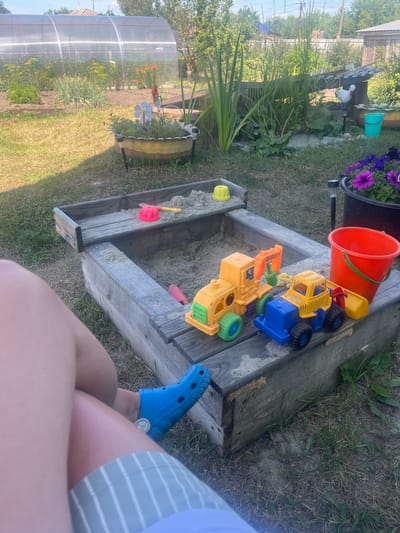 This media file shows a person's legs and feet, clad in blue Crocs and striped shorts, resting near a wooden sandbox filled with sand and toys. The sandbox contains several brightly colored plastic toys, including a yellow dump truck, a blue truck, and a red shovel. An orange bucket is also visible near the sandbox. In the background, lush green foliage and trees suggest an outdoor setting. To the left, a portion of a greenhouse can be seen, along with potted plants, one with vibrant purple flowers. The overall scene depicts a sunny day, likely in the afternoon, given the natural light. The setting appears to be a residential backyard or garden area. There is no visible text in the image.