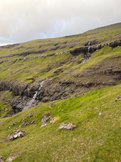 This is a wide, eye-level shot of a grassy, rocky hillside in Tjørnuvík, Faroe Islands, under a cloudy sky. The hillside is characterized by horizontal layers of dark rock interspersed with green grass. Several small waterfalls cascade down the rocky layers, flowing over the textured surface. The foreground is dominated by a gently sloping grassy area with scattered rocks of varying sizes, some covered in moss. The overall impression is one of a rugged, natural landscape.