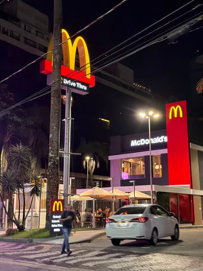 A McDonald's restaurant at night features a large sign with the golden arches and "Drive Thru." A white car is parked nearby. The restaurant building has outdoor seating with umbrellas. A man walks past a smaller McDonald's sign that reads "Entrada Estacionamento Drive Thru." Palm trees and other greenery are visible. Overhead power lines are present.