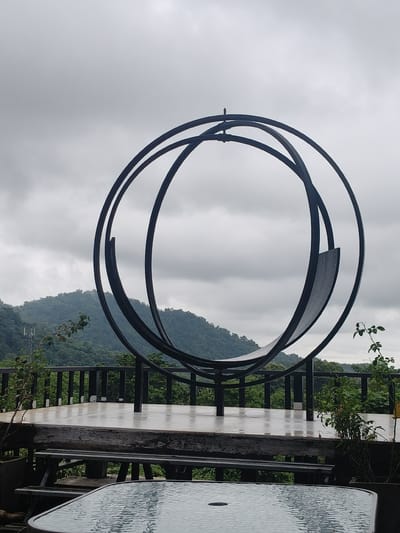 The image shows a large, dark, circular sculpture on a raised platform, possibly an outdoor seating area or a viewing deck. The sculpture appears to be made of metal and consists of several overlapping, curved bands. In the background, lush green mountains rise under a cloudy, overcast sky. A railing runs along the edge of the platform, and some vegetation, including a small tree with branches, is visible on the right side of the frame. In the foreground, a glass-topped table with a metallic frame is partially visible. The overall impression is of a serene, natural setting, potentially a tourist spot or a quiet retreat. While the location is stated to be Ban Pu Khai, Thailand, the image itself does not contain specific visual cues that would confirm this geographic detail.