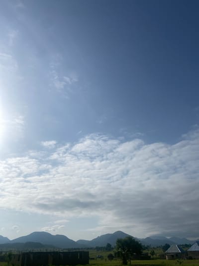 The image displays a landscape scene with a focus on the sky and mountains. The sky is predominantly blue with scattered white clouds. Towards the right side of the image, a range of mountains is visible, appearing hazy and silhouetted against the sky. In the foreground on the right, there is a grassy area with some buildings. The buildings appear to be under construction or in a state of disrepair, with some walls and roofs visible. A few trees are also scattered throughout the landscape. The overall scene suggests an outdoor setting in a rural or semi-rural area. The lighting indicates it is daytime, possibly late afternoon, given the soft light and the hazy appearance of the mountains. There is no visible text in the image.