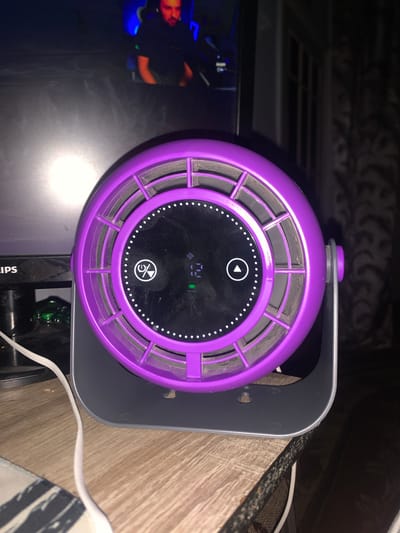 A purple fan with a digital display is on a wooden desk. The fan has a circular grill and touch controls on its front panel. Behind the fan, a computer monitor is visible, displaying a person sitting in a gaming chair. The environment appears to be indoors, with dim lighting. The image is shot from a medium distance, focusing on the fan and the monitor. The location is specified as Glodeni, Moldova.