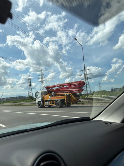 The image, oriented vertically, shows a street scene from inside a vehicle. A concrete pump truck with a long red boom is the primary object, positioned on the right side of the frame, traveling on an asphalt road. The truck is yellow and white, with visible wheels and some lettering on its side, though not clearly legible. The road has white lane markings.

In the background, large metal electricity pylons with numerous wires stretch across the frame, indicating an area with significant power infrastructure. The sky is a bright blue, filled with large, fluffy white clouds, suggesting fair weather. There are also buildings visible in the distance on the right side, forming a skyline. A street lamp is visible on the left side, positioned above the truck. The interior of the vehicle is partially visible on the right, including a dark gray dashboard area and part of a circular vent. The overall impression is of a daytime scene, possibly in a semi-urban or industrial area, during good weather. The context provided places the scene in Poroshkino, Russia.