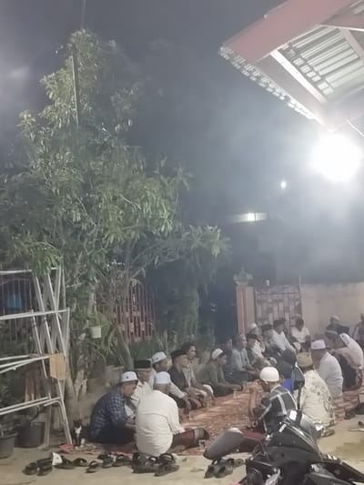 In Keudematangkuli, Indonesia, at night, a group of men are gathered on the ground, seated on a patterned rug. They are wearing traditional clothing, including long robes and skullcaps. Some of the men are engaged in conversation, while others appear to be listening. A motorcycle is parked in the foreground, and a small black and white cat is visible near a metal rack. The setting is outdoors, with trees and fencing visible in the background, suggesting a residential or communal area. The lighting suggests artificial illumination, possibly from a nearby building or a street light.