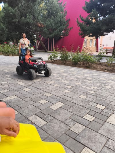 In Bayet, Kyrgyzstan, a young girl in an orange shirt drives a red toy car on a paved pathway. Behind her, a woman in a floral shirt and blue skirt stands near a rose bush. The background features trees, a building with a red corrugated metal facade, and a brick structure. The pathway is made of gray and cream colored rectangular pavers. In the foreground, a person's hand with a ring is visible resting on a bright yellow fabric.