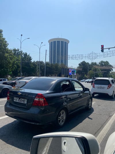 The image captures a street scene in Andijan City, Uzbekistan, under a clear blue sky. In the foreground, a dark gray Chevrolet Nexia LTZ sedan with license plate 60F 658HB is stationary in traffic. To its right, a white Chevrolet Spark with license plate 60H 387KB is also stopped. In the background, a distinctive cylindrical building with blue-tinted windows stands tall, flanked by trees and streetlights. A pedestrian crossing sign and a traffic light showing a red arrow are visible. Decorative string lights are suspended across the road. The perspective suggests the photo was taken from inside another vehicle.
