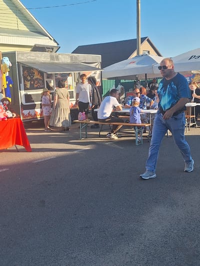 The image depicts an outdoor scene at what appears to be a local market or festival in the city of Tsіmkavіtskі sel'skі Save't, Belarus. The foreground is dominated by an asphalt surface with some white painted lines. A man in a blue t-shirt with a wolf graphic and light blue jeans is walking towards the viewer from the right side of the frame. He is wearing sunglasses and grey sneakers.
In the midground, several people are gathered around tables and a food truck. A food truck with a flame graphic and a menu displayed is visible on the left. To the right of the food truck, a woman in a long, tiered dress is standing and talking to a young girl in a patterned dress and a straw hat. Further to the right, a group of people are seated at tables under white umbrellas. One person is wearing a white t-shirt, and another is wearing a patterned shirt. A young child in a purple outfit is sitting on a bench at one of the tables.
In the background, buildings with light-colored siding and dark roofs are visible, along with utility poles and streetlights. The sky is clear and blue, suggesting a daytime setting. A red table cloth covers a table on the far left, displaying stuffed toys. The overall scene suggests a casual, communal gathering.
