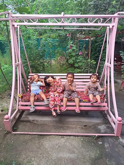 A woman and three children are sitting on a pink metal swing set with a patterned cushion. The woman, in the center, is wearing a floral print dress and has her eyes closed and head tilted back. To her left, a young boy in a light blue t-shirt and blue shorts is sitting with his legs outstretched, making a "peace" sign with his fingers. To the woman's right, two young girls are seated. The girl closest to the woman is wearing a brown t-shirt and leopard-print shorts, with her hand on her chin. The girl furthest to the right is also wearing a brown t-shirt and leopard-print shorts, and is holding onto the swing's frame. The swing is situated outdoors in a garden or yard, with lush green foliage and trees in the background. A green fence is partially visible behind the swing. The ground beneath the swing appears to be concrete and dirt. The lighting suggests it is daytime. The overall scene conveys a relaxed and playful atmosphere. There is no visible text in the image.