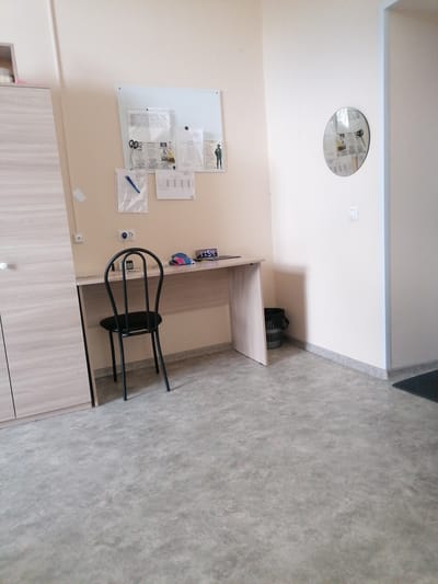 The image depicts a sparsely furnished room, likely a dormitory or small apartment, in Komosomolskiy, Russia. The room contains a desk with a black metal chair, a digital clock displaying "11:54", a small calculator, and a few personal items. A notice board with various papers and documents is mounted on the wall above the desk. To the left, a tall wooden wardrobe is visible, and to the right, a round mirror hangs on the wall. A small waste bin is situated next to the desk. The floor is covered with a grey, textured material, and the walls are painted a light, neutral color. The overall impression is one of functional simplicity.