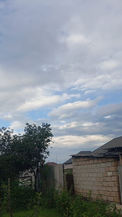The image depicts a cloudy sky over a rural scene, likely in Baksan, Russia. The sky is filled with a mix of gray and white clouds, with patches of blue visible in between.

In the foreground, to the left, is a lush green tree with dense foliage. Below the tree, there is an abundance of green bushes and some smaller plants. To the right of the tree, there are various structures, including a corrugated metal fence, a concrete wall, and a brick building. The brick building has a dark, peaked roof. Further in the background, more houses and roofs are visible, suggesting a residential area.

The overall impression is of an overcast day in a semi-rural or suburban setting. The vegetation is healthy and green, indicating a potentially warm season. The light suggests it could be late afternoon or early morning, given the diffused light from the clouds. There are no people visible in the image, and no text is present. The scene evokes a sense of quietude and natural ambiance.