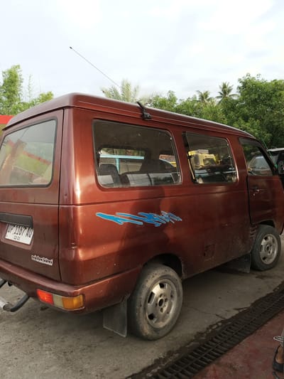 A dark red van is parked outdoors on a paved surface. The van has multiple windows, including rear passenger windows and a driver's side window. The van has a license plate that reads "DP 1167 AQ" and a sticker below it that says "mister king". There is a blue decal on the side of the van. The van appears to be dusty and worn. In the background, there are trees and a cloudy sky. The scene appears to be a street or parking area in Tocina, Indonesia.