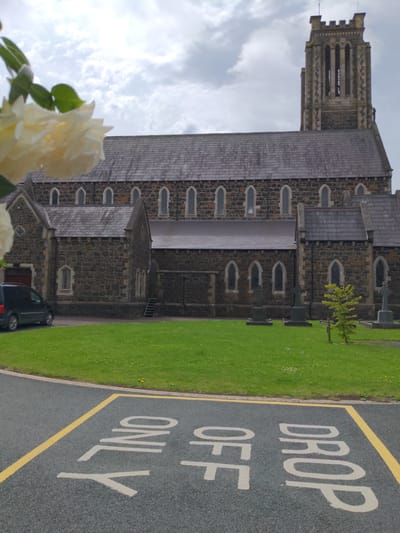 The image shows a stone church building with a tall tower, possibly a cathedral or a large parish church, situated in Ballymena, United Kingdom. The building has a grey tiled roof and arched windows. To the right of the church, there is a well-maintained green lawn with some small trees and what appear to be gravestones. In the foreground, to the left, a cluster of light-colored roses is visible, partially obscuring the view of the church.
The ground in front of the church is paved, and a section of this paving has markings indicating a "DROP OFF ONLY" zone. The text is painted in large, light-colored letters on the dark asphalt. A yellow line borders the marked area. A dark-colored car is parked in the vicinity of the drop-off zone.
The sky is overcast with clouds, suggesting a possibly dull or rainy day. The image is oriented vertically, with the top of the church tower pointing upwards. The scene appears to be a quiet, possibly solemn or peaceful setting, given the presence of a church and gravestones, yet the "DROP OFF ONLY" sign suggests it is also a functional area, perhaps for a school or community center associated with the church. There are no people visible in the image. The time of day is indeterminate due to the overcast conditions.