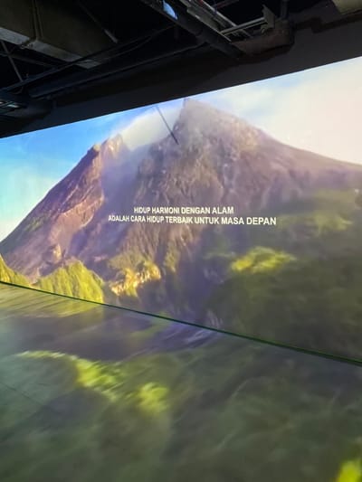 The image shows a projection of a mountain range on a wall in a room. The text on the screen reads "HIDUP HARMONI DENGAN ALAM ADALAH CARA HIDUP TERBAIK UNTUK MASA DEPAN". The projection is projected from a projector mounted to the ceiling. The room is empty and the walls are grey. The floor is covered in the reflected image of the projected mountain range. The coordinates -7.8 latitude and 110.407 longitude place this location in Banguntapan, Indonesia. The projection appears to be part of an educational or artistic installation.